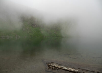 Foto: ein See mit ruhigem Wasser und vorbeiziehenden Nebelschwaden, im Vordergund eine Schieferplatte